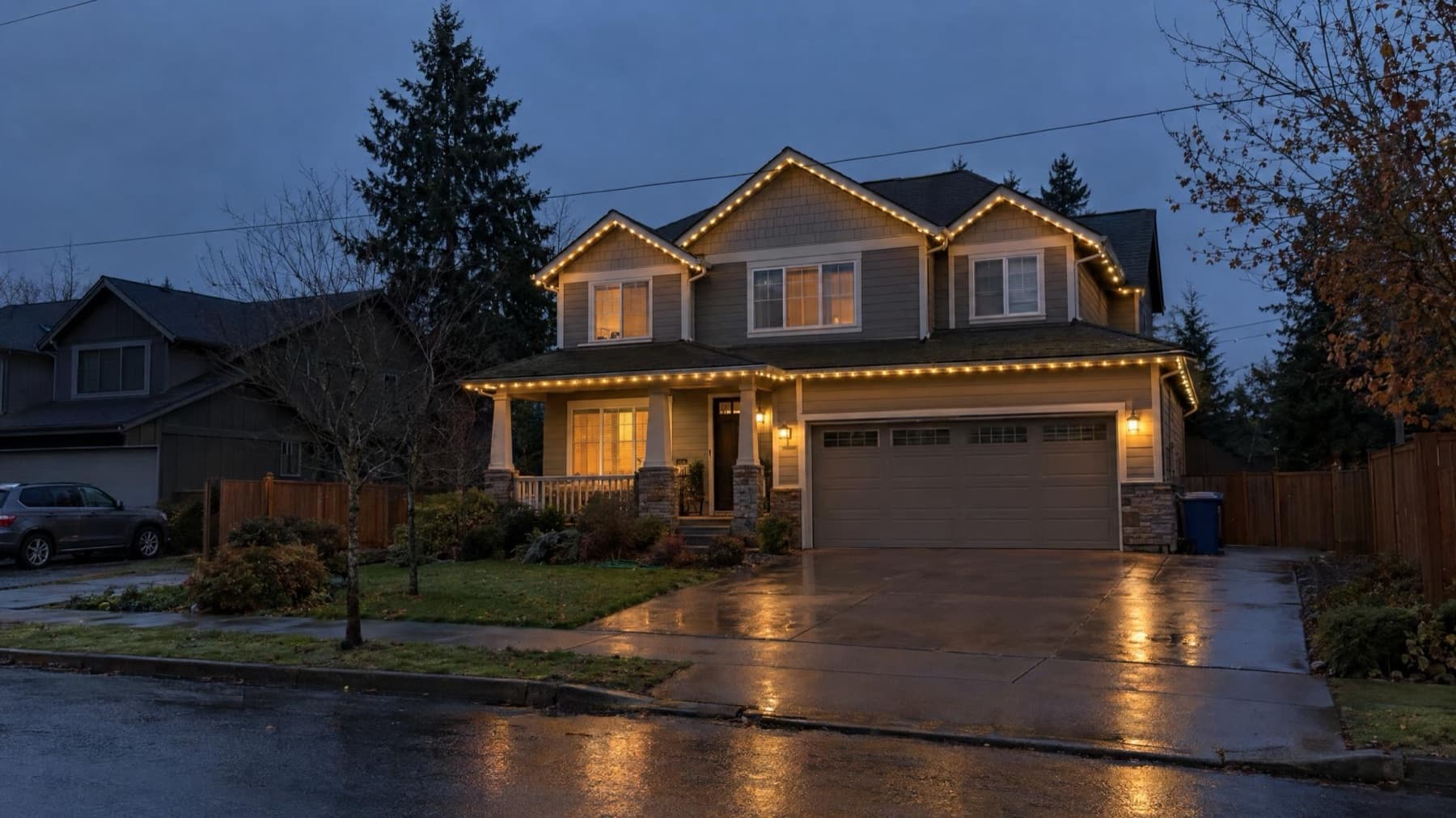 Two-story craftsman at dusk with a warm-white lit roofline, wet street in the foreground