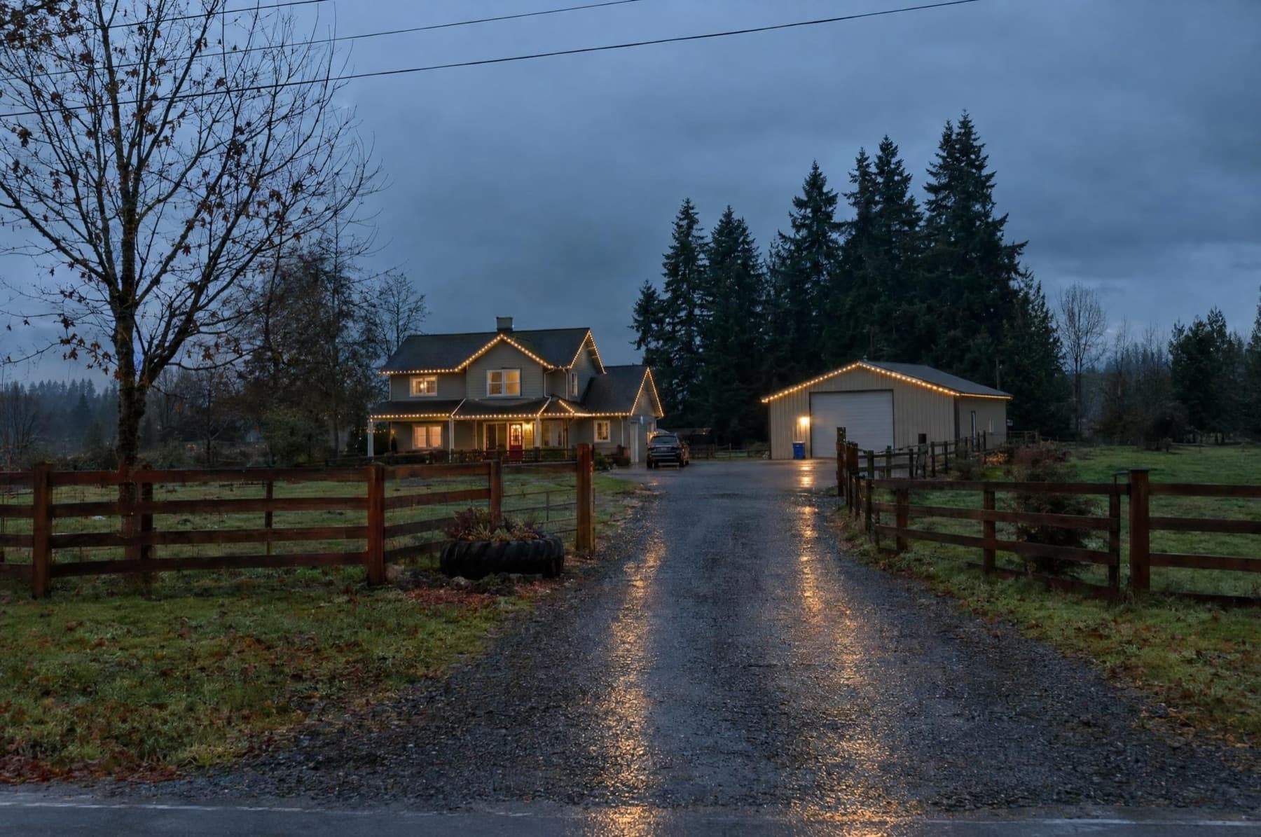 Farmhouse and steel shop both lit warm white at the end of a gravel driveway